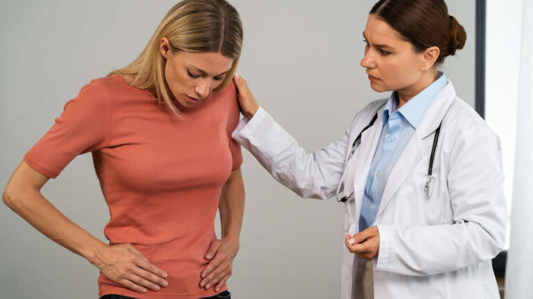 A woman sitting at her office desk, discreetly holding her lower abdomen, showing the disruptive effect of chronic pelvic pain.